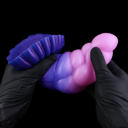 Larval the Alien Spawn dildo, Flux colorway, flexed between two gloved hands to show the silicone's give